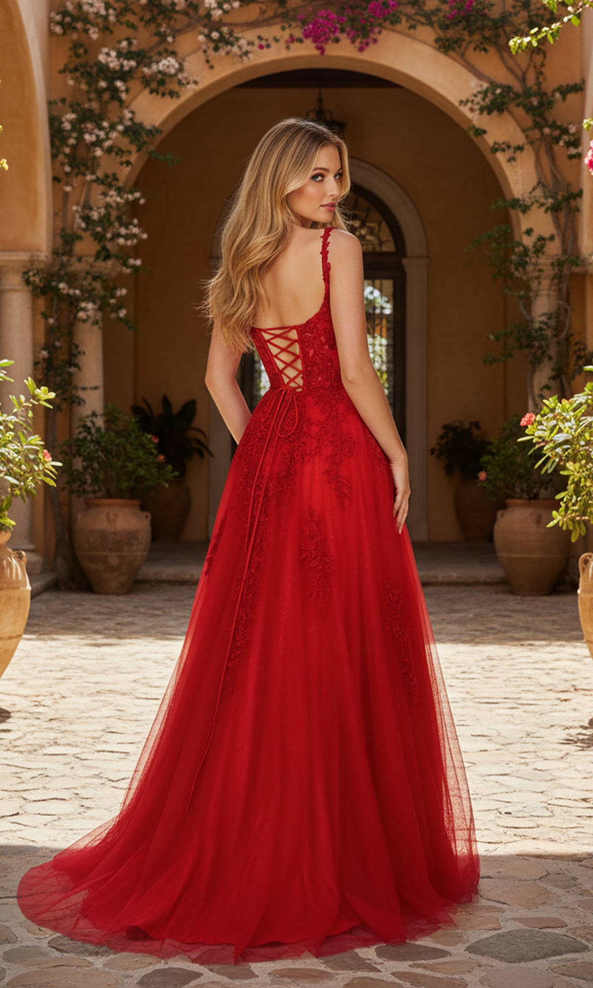 A woman wearing the PromGirl Exclusive 250233 long prom dress stands outdoors on a stone patio, facing away, with greenery and an arched doorway in the background.